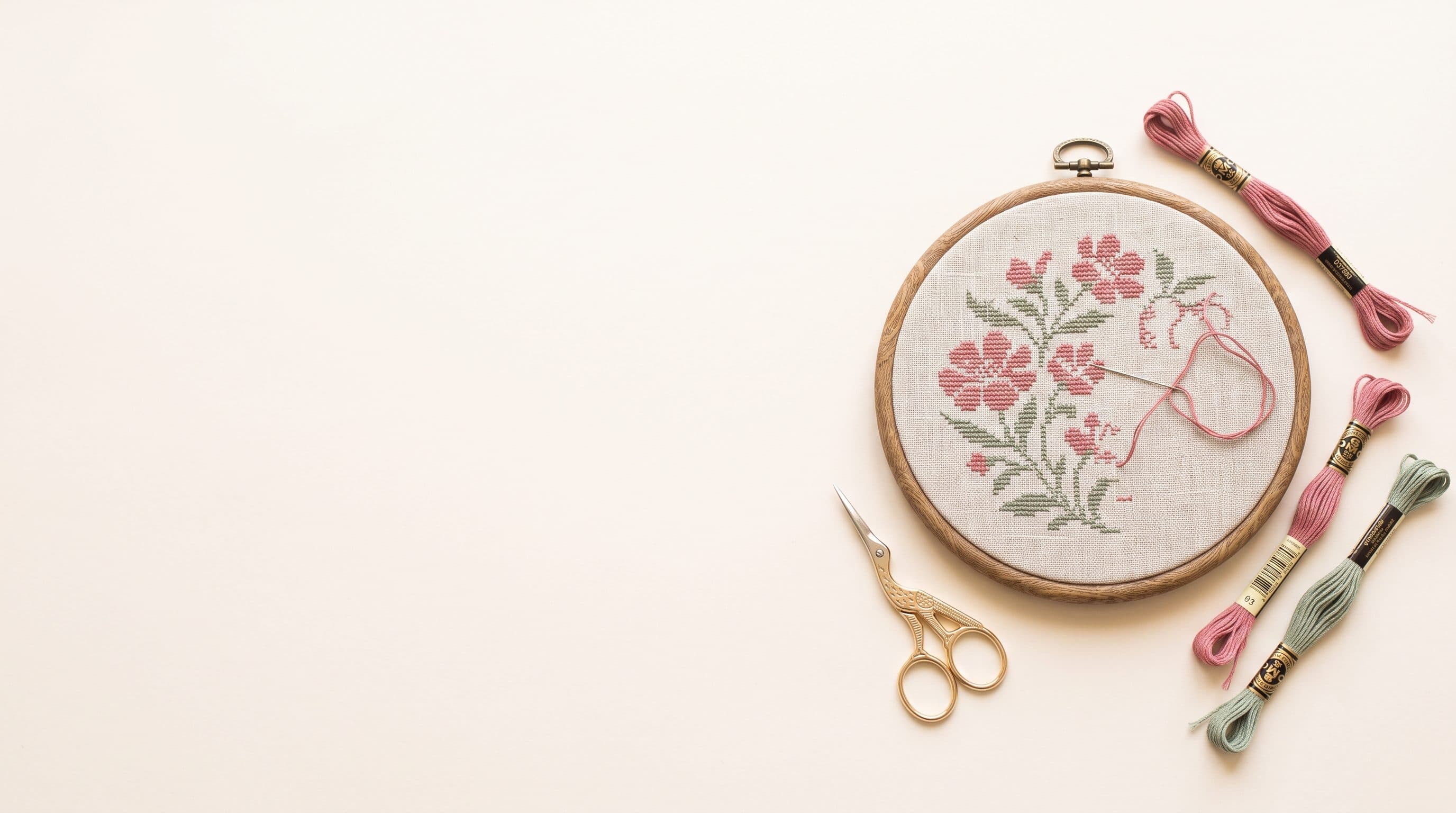 A part-finished cross stitch in a wooden embroidery hoop, surrounded by skeins of coloured floss